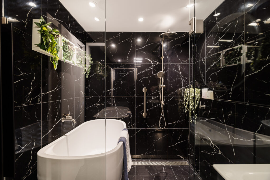 Renovated Bathroom, with black gloss tiles on walls, bath sitting on the side with shower and grab rail adjacent.